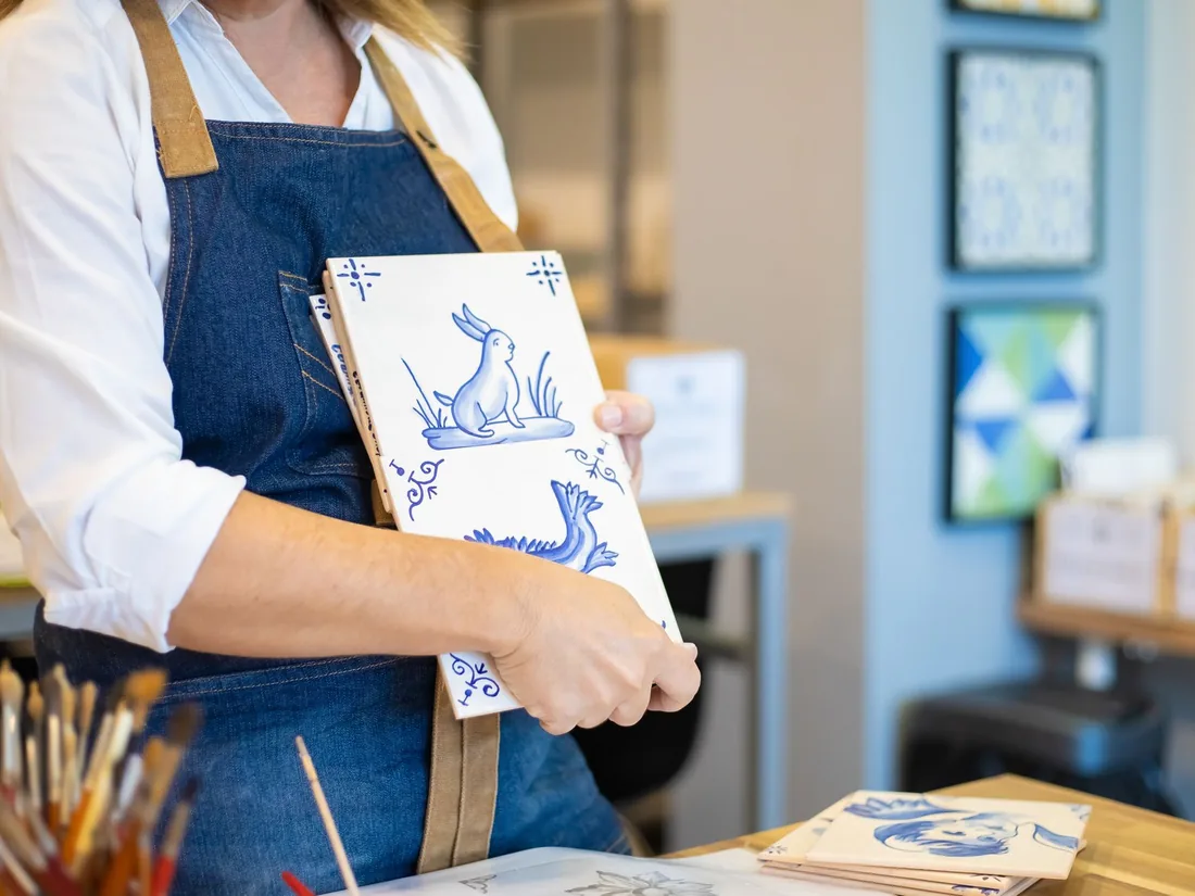 Azulejos pintados a mano en azul cobalto sostenidos sobre el delantal en un taller de cerámica, con pinceles sobre la mesa